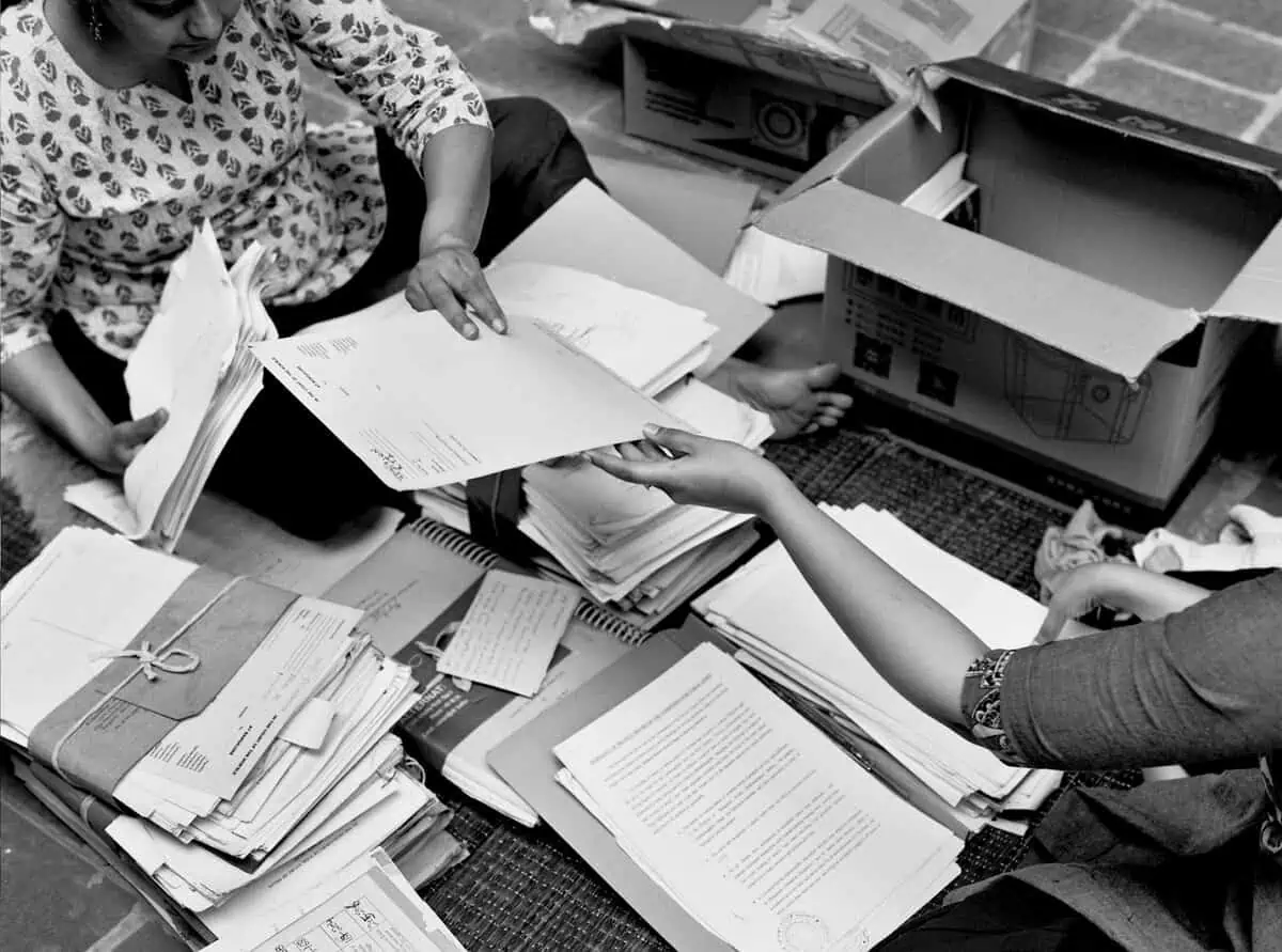 Two women sitting on the floor, sorting through stacks of papers and files from cardboard boxes.